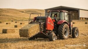 A red tractor operates a large square baler making tight golden hay bales on a different modern hay farm.