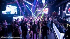 Realistic view of a bustling modern nightclub interior with a DJ, crowded dance floor, laser lights, and illuminated bar area.