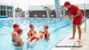 Lifeguard trainees in red uniforms practice a group dummy rescue in a bright, modern outdoor swimming pool.