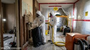 Two technicians in hazmat suits removing contaminated drywall inside a sealed residential asbestos abatement area.