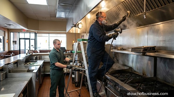 Two technicians clean the large stainless steel exhaust hood in a commercial kitchen.